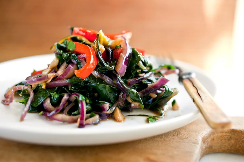 A plate of stir-fried Swiss chard with red and yellow bell peppers and red onions, garnished with a wooden handled fork on the side, on a light-colored table.