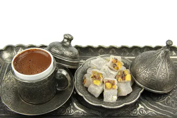 A traditional Turkish coffee set with a metal cup and saucer with intricate patterns, accompanied by a metal sugar bowl with a lid, and a plate of Turkish delight topped with pistachios, arranged on an ornate metal tray, reflecting the customary serving of Turkish coffee.