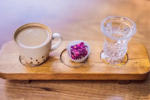 A modern presentation of coffee on a wooden board, featuring a white porcelain cup, a glass of water, and a single piece of Turkish delight in a paper cup, showcasing a contemporary twist on serving Turkish coffee.