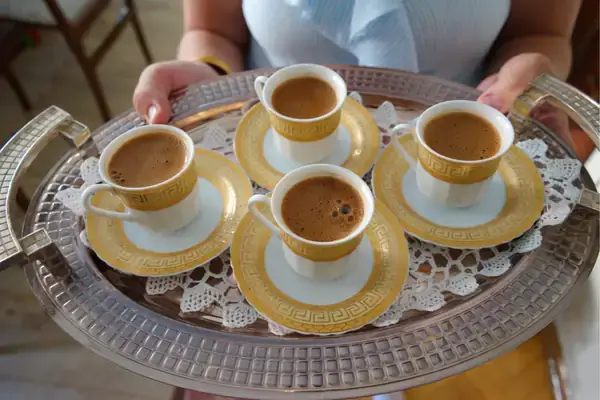 A woman's hands holding a silver tray adorned with lace, serving four porcelain cups of Turkish coffee with golden accents, capturing the communal aspect of sharing coffee.