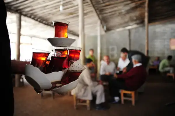 A blurred background of people sitting in a traditional setting, with a focused foreground of a hand carrying a tray of tea glasses stacked in a pyramid, illustrating the communal and generous spirit of tea serving in Turkish social gatherings.