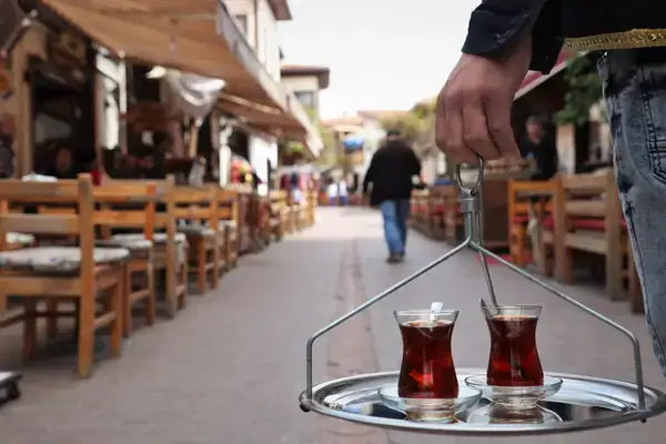 A close-up shot capturing a person's hand carrying a metal tray with two traditional glasses of Turkish tea. The tray is being served against a blurred streetscape background, depicting a bustling pedestrian lane lined with wooden tables and chairs outside local eateries, evoking the daily life and culture of street-side tea drinking in Turkey