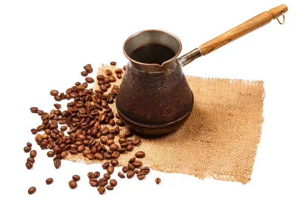 A cezve, a traditional Turkish coffee pot with an ornate design and a wooden handle, lies on a burlap fabric surrounded by scattered coffee beans, suggesting the process of making fresh Turkish coffee