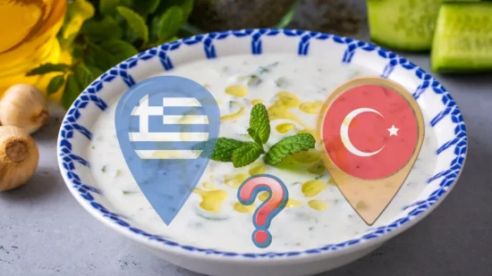 This image showcases a dish that closely resembles both Greek Tzatziki and Turkish Cacık, served in an ornate white bowl with a blue pattern. The dish consists of a creamy yogurt base dotted with slices of fresh cucumber, a drizzle of golden olive oil, and a sprinkle of mint leaves for garnish. Ingredients such as whole cucumbers and bulbs of garlic are visible in the background, indicating their use in the recipe. The dish is set against a dark, textured surface, highlighting the bright, appetizing colors of the food.