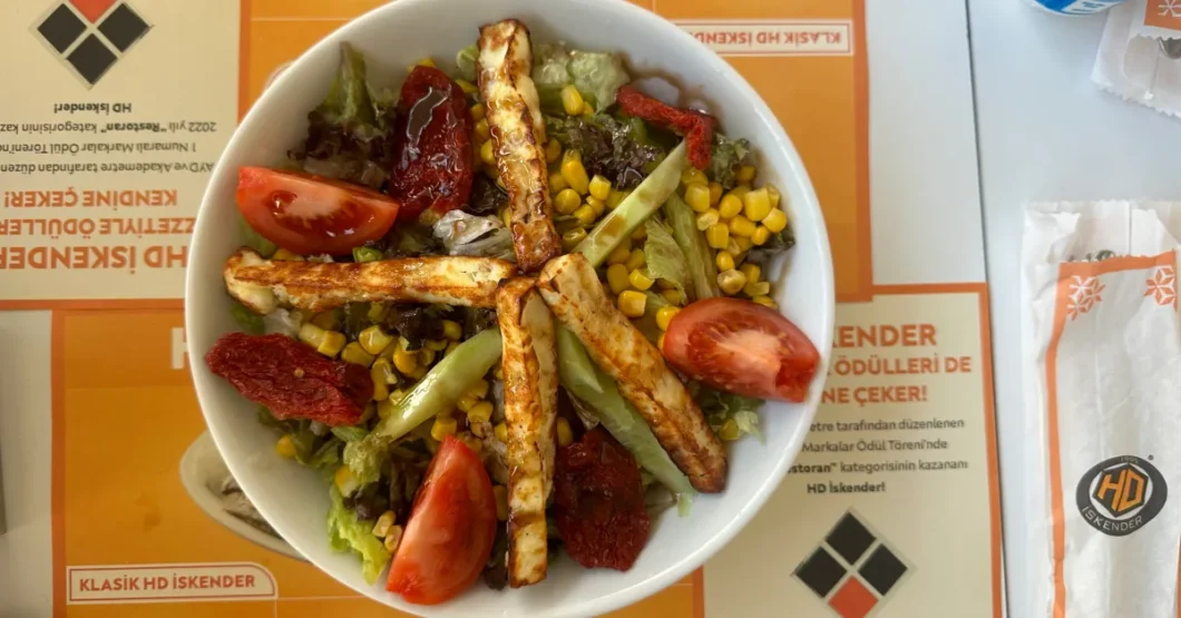 the salad is viewed from above, giving a clear overview of the dish's composition. The grilled halloumi cheese strips are arranged in a crisscross pattern, and the salad ingredients are more evenly distributed, offering a balanced mix of colors and textures. The background includes a part of the restaurant's menu and a wrapped sandwich, hinting at more offerings from the eatery.