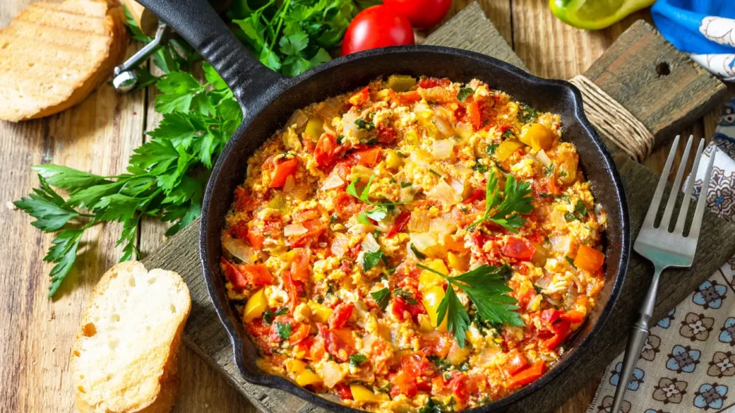 The first image displays a close-up of menemen, a traditional Turkish dish, served in a black cast-iron skillet. The skillet sits on a wooden board, accompanied by slices of bread, a fresh tomato, and a sprig of parsley. The menemen itself is colorful, with bright red tomatoes, green peppers, and yellow eggs mixed together, garnished with green parsley on top.