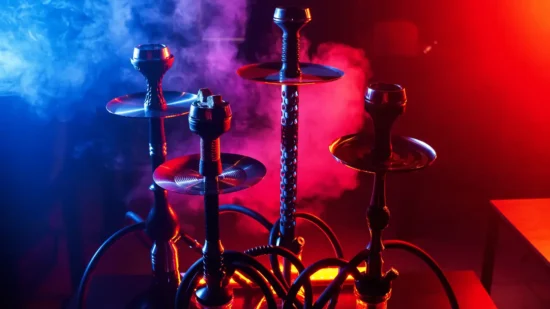 An atmospheric image of multiple hookahs lined up against a dark background with moody lighting emphasizing red and blue hues. The hookahs have tall stems and transparent bases through which smoke is visible, giving off a vibrant and alluring vibe.
