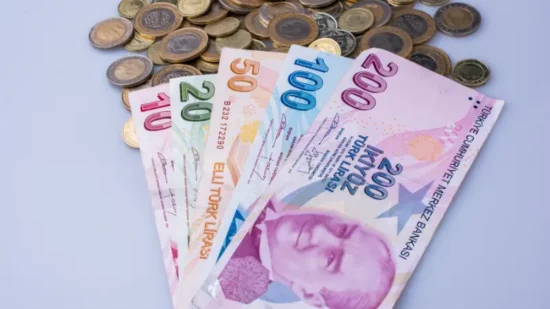 Turkish lira banknotes in denominations of 20, 50, 100, and 200 are fanned out on a surface, with an assortment of coins, both Turkish and various others, scattered behind them.