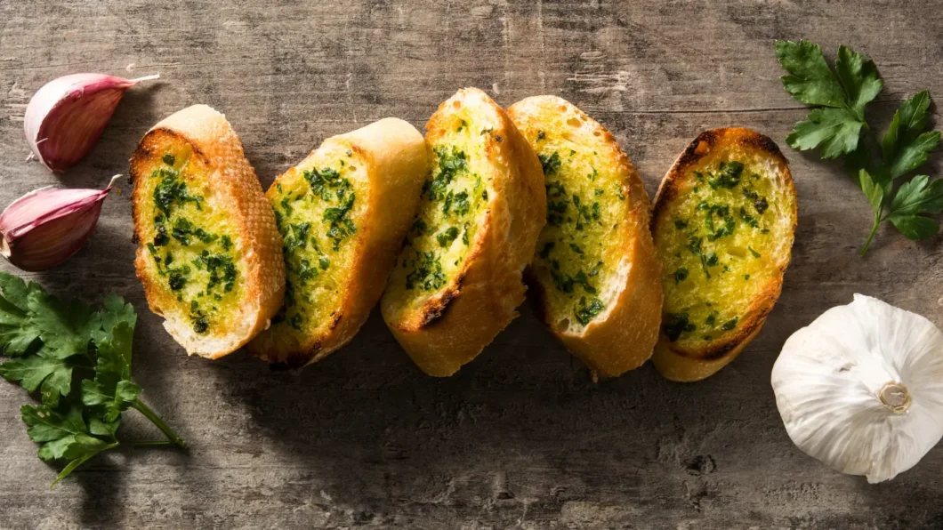slices of garlic bread arranged on a rustic wooden surface, with cloves of garlic and sprigs of parsley scattered around to enhance the composition. The bread boasts a golden-brown crust, with the garlic and parsley mixture visible on its toasted surface, suggesting it's just been baked following a garlic bread recipe.