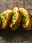 slices of garlic bread arranged on a rustic wooden surface, with cloves of garlic and sprigs of parsley scattered around to enhance the composition. The bread boasts a golden-brown crust, with the garlic and parsley mixture visible on its toasted surface, suggesting it's just been baked following a garlic bread recipe.