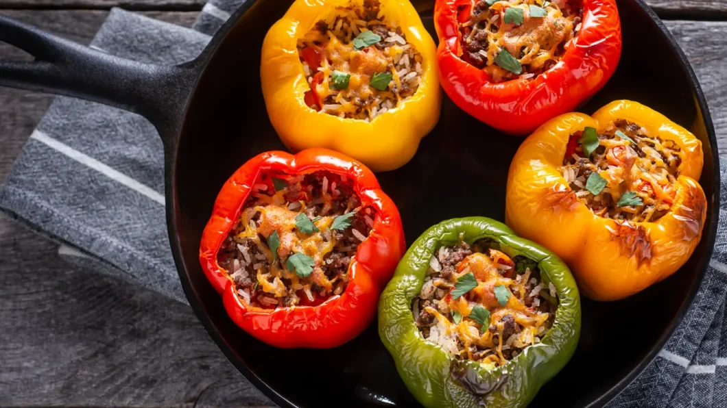 Four bell peppers, vibrant red and yellow, are stuffed with a savory mixture of cooked rice and herbs, all lovingly prepared with olive oil. They're beautifully presented in a black cast iron skillet, as the epitome of the dish known as stuffed peppers with olive oil, ready to be served.