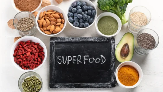 array of superfoods arranged neatly around a chalkboard with the word "SUPERFOOD" written on it. This spread includes bowls of almonds, blueberries, goji berries, broccoli, turmeric, and other healthy ingredients like chia and flax seeds.