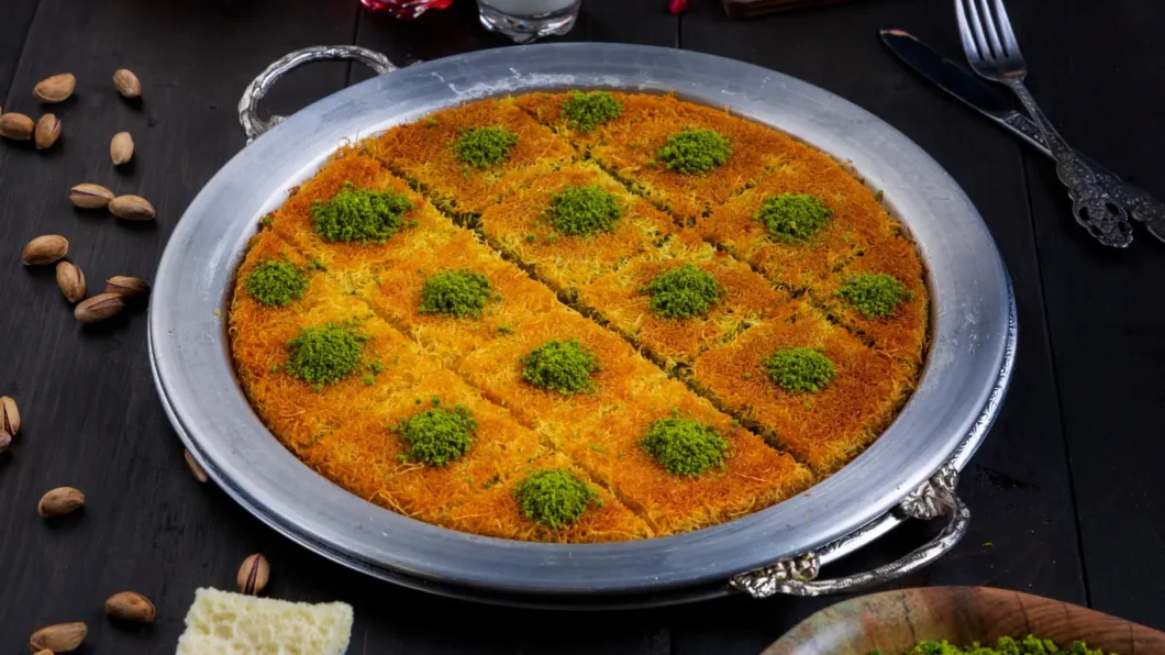 A large, completed kunefe is presented on an ornate silver tray, cut into even triangular portions, each topped with a vibrant green sprinkle of ground pistachios, contrasting with the bright orange strands of cooked kadayıf. The dessert is set against a dark background, accentuating its rich colors and inviting texture.
