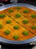 A large, completed kunefe is presented on an ornate silver tray, cut into even triangular portions, each topped with a vibrant green sprinkle of ground pistachios, contrasting with the bright orange strands of cooked kadayıf. The dessert is set against a dark background, accentuating its rich colors and inviting texture.