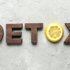 features the word "DETOX" laid out in bold, dark wooden letters on a textured light grey concrete background. A bright, fresh slice of lemon is placed next to the letter "X," adding a touch of color and suggesting a connection to natural, detoxifying foods.