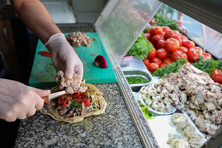 a street food vendor preparing a wrap of kelle söğüş. The vendor, wearing gloves, is placing slices of the cooked meat onto a flatbread, which is already garnished with chopped herbs, onions, and tomato pieces. This scene takes place at an outdoor food stall, with ingredients like tomatoes and greens visible in the background.