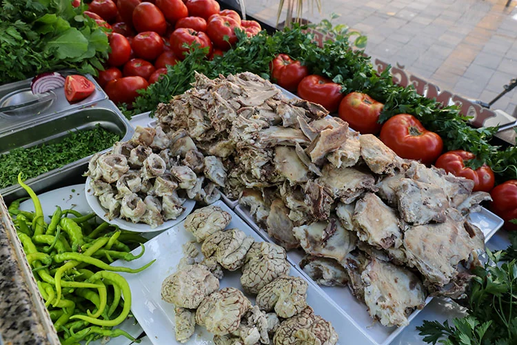image shows a closer look at the ingredients available at the street food stall. There is a large pile of cooked sheep's head pieces in the center, surrounded by smaller plates containing specific parts like brains and cheeks. Fresh vegetables, including tomatoes, green peppers, and bunches of parsley, are neatly arranged around the meat, highlighting the freshness and variety used in preparing this traditional dish.